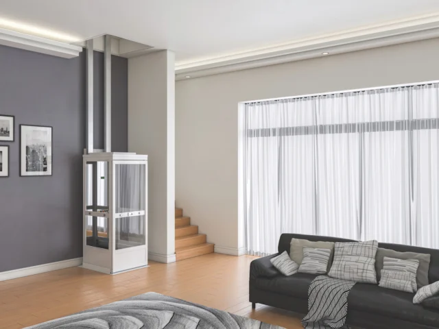 Modern living room with a small glass residential elevator near wooden stairs, gray sofa with cushions, and large window with sheer curtains.