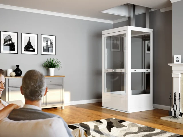 A couple sits on a sofa in a modern living room with a white residential elevator, wall art, fireplace, and hardwood floors.