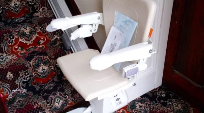 A beige stairlift chair is installed on a staircase with ornate red and blue patterned carpet and white paneled walls.