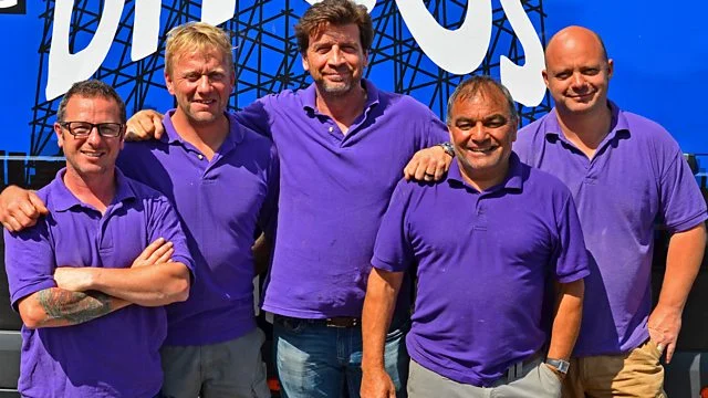 Five men wearing purple polo shirts stand side by side outdoors in front of a blue and white sign, smiling at the camera.