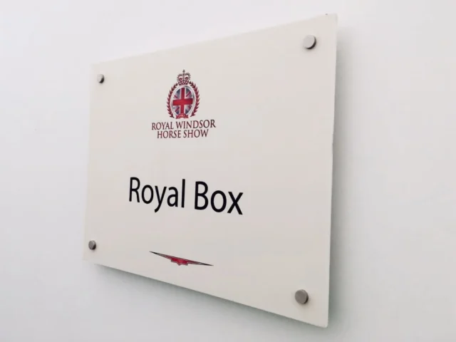 White sign with the Royal Windsor Horse Show logo and text that reads "Royal Box" mounted on a white wall with four visible screws.