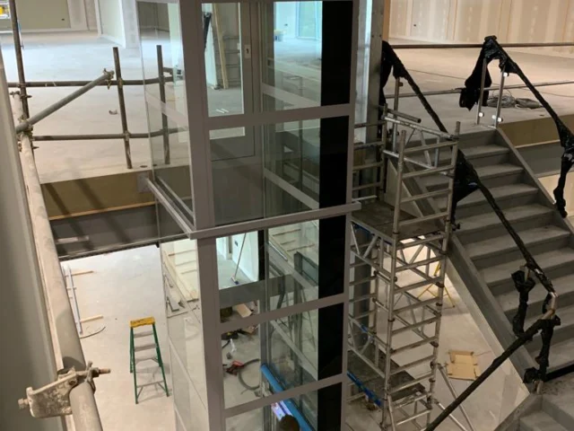 A glass elevator under construction is installed between two staircases in a building, surrounded by scaffolding and construction materials.