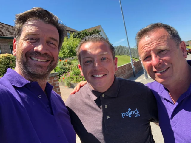 Three men are standing outside on a sunny day, smiling at the camera. Two wear purple shirts and one wears a dark shirt with a "Pollock Lifts" logo.