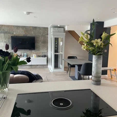 Modern open-plan living space with a kitchen island, dining table, flowers in vases, wall-mounted TV, and a small residential elevator in the corner.