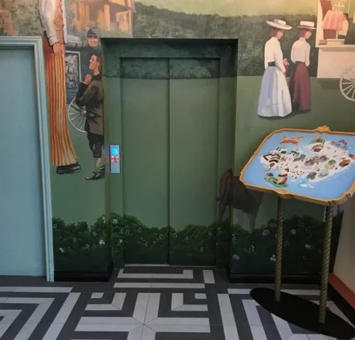 An elevator door painted to blend in with a mural depicting people in vintage clothing, next to a map on a stand and patterned floor tiles.
