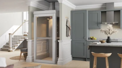 A small residential elevator installed in a modern kitchen area, with gray cabinets, a staircase, and bar stools visible nearby.