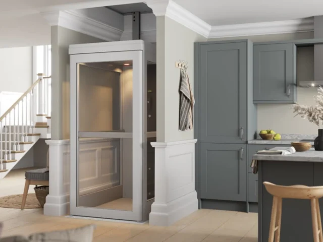 A small residential elevator installed in a modern kitchen area, with gray cabinets, a staircase, and bar stools visible nearby.