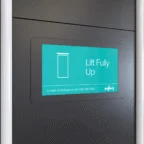 A digital display on a dark wall shows a door icon and the text "Lift Fully Up" with an emergency contact number below.