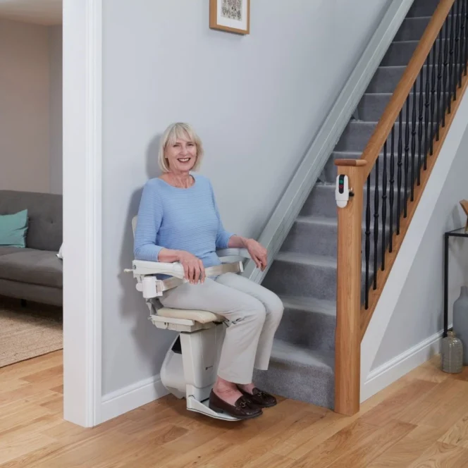An older woman sits on a stairlift positioned at the base of a carpeted staircase in a modern, well-lit living room.