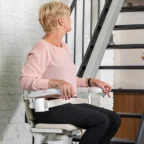 An older woman sits on a stairlift at the base of a staircase, looking to her left in a modern home interior.