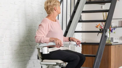 An older woman sits on a stairlift at the base of a staircase, looking to her left in a modern home interior.