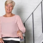 Older woman with short blonde hair sits on a stairlift by a white brick wall and staircase, holding the control and smiling at the camera.