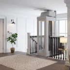 A modern residential elevator with glass panels is installed next to a staircase in a bright, carpeted hallway with white walls and black railings.