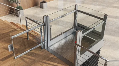 A modern indoor wheelchair platform lift with glass panels is installed beside a staircase in a building lobby with elevators and potted plants.