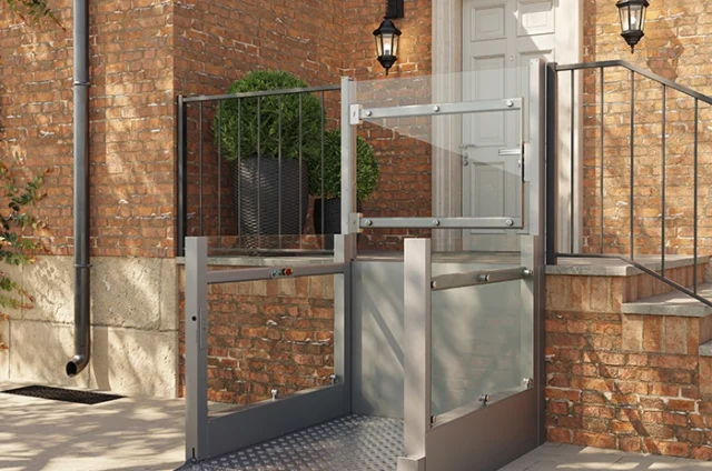 A metal wheelchair lift installed outside a brick building provides accessible entry to a raised doorway with stairs and black railings.