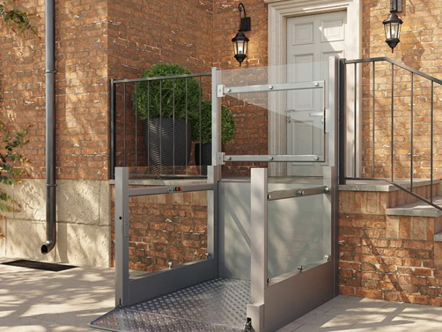 A metal wheelchair lift installed outside a brick building provides accessible entry to a raised doorway with stairs and black railings.