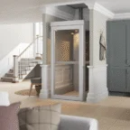 A modern residential elevator installed in a living room, with light wood flooring, a staircase, and gray cabinetry visible in the background.