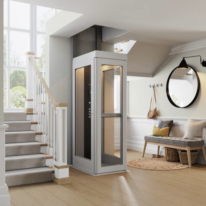 A compact residential elevator is installed beside a staircase in a modern, well-lit home entryway with a bench, mirror, and coat hooks.