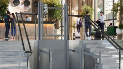 A modern hotel lobby with marble floors, a front desk, several people, and a platform lift for wheelchair accessibility between two staircases.