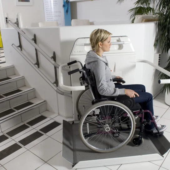 A woman in a wheelchair uses a platform lift beside a staircase in a modern indoor setting with white tiles and a large plant.