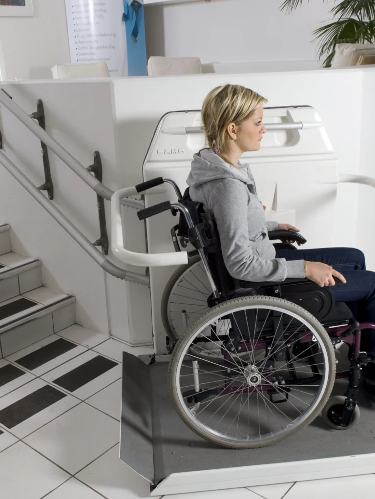 A woman in a wheelchair uses a platform lift beside a staircase in a modern indoor setting with white tiles and a large plant.