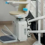 A motorized stair lift with a beige seat and blue armrest is installed on a curved track along a staircase in a modern home interior.