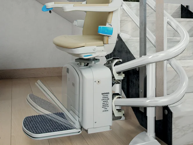 A motorized stair lift with a beige seat and blue armrest is installed on a curved track along a staircase in a modern home interior.