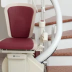 A motorized stair lift with a maroon seat and control panel is installed beside a curved staircase with brown steps.
