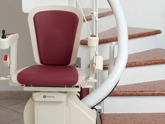 A motorized stair lift with a maroon seat and control panel is installed beside a curved staircase with brown steps.