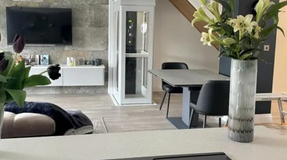 Modern open-plan living space with a kitchen island, dining table, wall-mounted TV, and a home lift in the background; vases of flowers decorate the room.