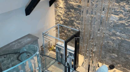A person works near a glass elevator and a tall, hanging light fixture inside a modern room with stone walls, marble floors, and a skylight.