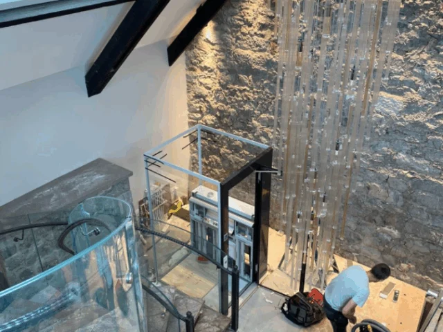 A person works near a glass elevator and a tall, hanging light fixture inside a modern room with stone walls, marble floors, and a skylight.