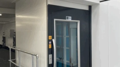 A modern elevator with a glass window and dark frame is situated in a clean, indoor industrial setting with safety railings and a fire extinguisher nearby.
