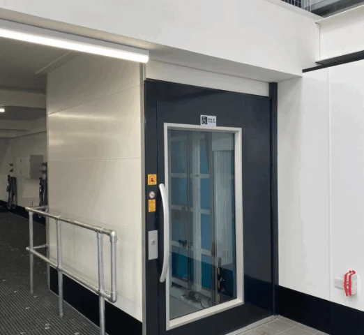 A modern elevator with a glass window and dark frame is situated in a clean, indoor industrial setting with safety railings and a fire extinguisher nearby.