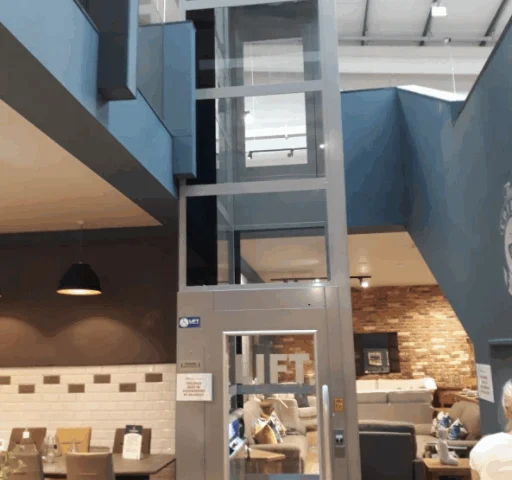 A small glass home elevator is installed indoors between two floors, surrounded by modern furniture and decor.