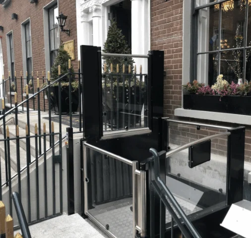 A wheelchair lift installed beside a stairway outside a brick building, providing accessible entry to the main door.