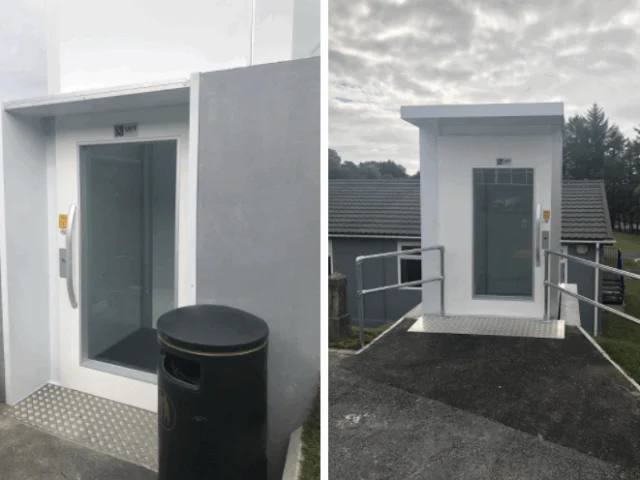 Outdoor elevator entrance with a glass door, metal ramp, and handrails, positioned next to a trash bin and surrounded by buildings and trees.