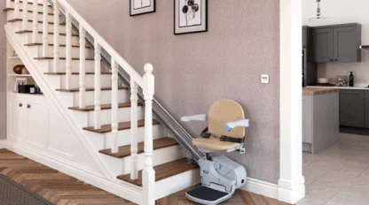 A stairlift chair is installed on the side of a wooden staircase in a modern home, with a kitchen visible in the background.