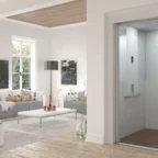 A modern living room with large windows and minimalist decor features a residential elevator with open doors next to a white wall.