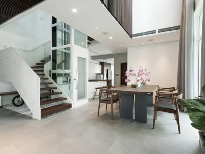 Modern open-plan interior with a staircase, glass elevator, dining table with chairs, potted plant, and minimalistic decor.