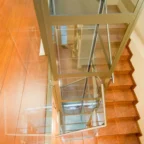 A glass elevator shaft is positioned next to a wooden staircase with brown steps in a well-lit interior space.