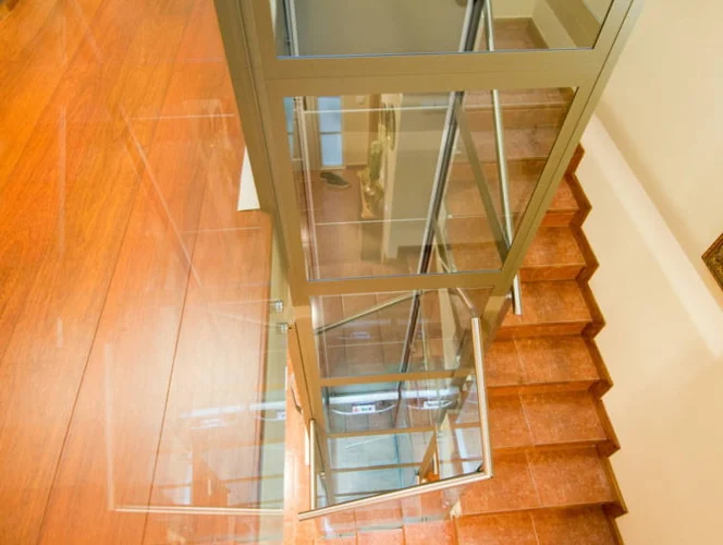 A glass elevator shaft is positioned next to a wooden staircase with brown steps in a well-lit interior space.