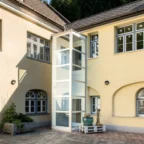 A modern glass elevator is installed on the exterior of a yellow two-story building with multiple white-framed windows and a paved courtyard.