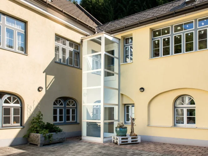 A modern glass elevator is installed on the exterior of a yellow two-story building with multiple white-framed windows and a paved courtyard.