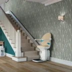 A beige stairlift installed along a carpeted staircase in a home with green patterned wallpaper and wooden flooring.