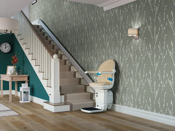 A beige stairlift installed along a carpeted staircase in a home with green patterned wallpaper and wooden flooring.