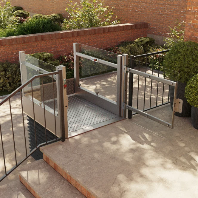 A small outdoor platform lift with glass sides and a metal gate, installed beside stairs for wheelchair accessibility. Shrubs and a brick wall are in the background.
