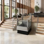 A modern indoor space features an accessibility lift between two sets of stairs, surrounded by glass panels and metal railings, with large windows and plants in the background.