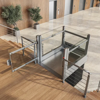 A modern wheelchair lift with glass panels is installed next to a staircase in an indoor public building with elevators and potted plants.