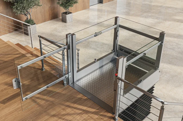 A modern wheelchair lift with glass panels is installed next to a staircase in an indoor public building with elevators and potted plants.